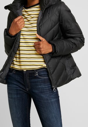 Person iført en sort quiltet jakke over en gul og hvid stribet skjorte og mørkeblå jeans, der justerer jakke lynlåsen.