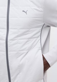 Veste matelassée blanche avec fermeture éclair grise et logo Puma sur la poitrine, avec une main à la peau foncée placée dans la poche du côté droit.