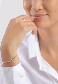 Silver bracelet with an infinity symbol and heart accents, featuring a delicate chain design. The wearer is in a white shirt, resting chin on hand.
