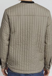 Quilted beige jacket with a textured finish, black collar, and buttoned cuffs. Features horizontal stitching patterns and a relaxed fit.