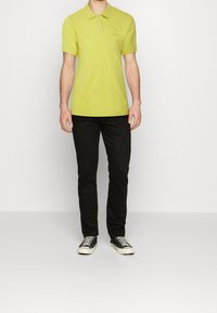 Polo vert citron avec col, manches courtes et un logo discret. Associé à un pantalon noir et des baskets noires à semelles blanches.
