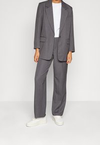 Blazer grigio su misura con revers oversize e due tasche frontali, abbinato a pantaloni larghi coordinati e sneakers bianche.