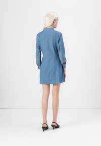 Denim shirt dress in light blue, with long sleeves and a fitted waist. Features a collared neckline and a mid-thigh length. Black heeled shoes.