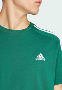 Zaļš kokvilnas T-krekls ar apaļu kakla izgriezumu, uz kura pleciem ir trīs baltas svītras un baltā Adidas logo uz krūšu daļas.