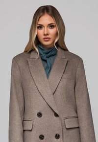 Ung kvinde med langt blond hår iført en grå dobbeltbrystet frakke og et tealfarvet tørklæde knyttet om halsen mod en ensfarvet baggrund.