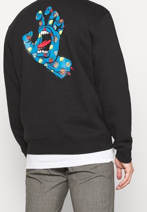 Blåt grafik-hånd med åben mund, tunge stikkende ud og farverige mønstre på ryggen af en sort sweatshirt, båret med grå bukser og lagdelt hvid skjorte.