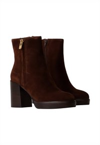 Botas de tobillo de ante marrón con un tacón grueso y bloque, y cremallera dorada. Textura suave y diseño minimalista sin acentos adicionales.