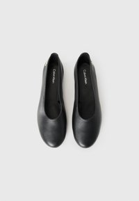 Een paar zwarte leren ballerina's van Calvin Klein met ronde neuzen, op een effen lichtgrijze achtergrond.