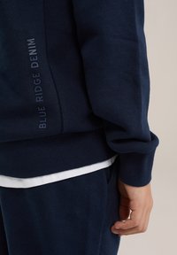 Navy pullover sweatshirt with ribbed cuffs and hem, featuring "BLUE RIDGE DENIM" logo in dark blue along the side, layered over a white shirt.