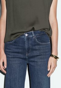 Ciemnoniebieskie dżinsy z prostym krojem, z przednimi kieszeniami, srebrnym guzikiem i beżowym przeszyciem. Noszone z oliwkowym topem.