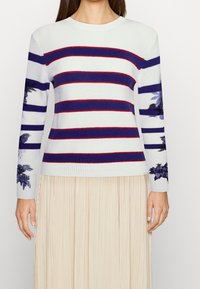 Striped sweater in white, navy, and red, featuring floral accents on sleeves. Ribbed texture with a crew neck and long sleeves.