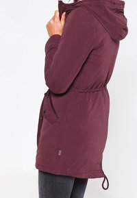 Burgundy huva jacka med dragsko i midjan, dragkedja och knappstängning, samt sidofickor. Slät textur och knälång design.