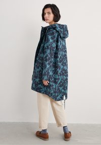 Waterdichte jas met een teal en marineblauw bloemenpatroon, capuchon, drukknoopsluiting en een vrije pasvorm, gecombineerd met lichtgekleurde broeken en bruine schoenen.