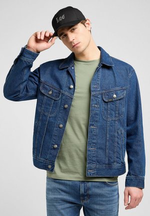Giacca di jeans blu scuro con chiusura a bottoni, due tasche sul petto e una vestibilità rilassata, indossata sopra una camicia verde chiaro e abbinata a jeans blu.
