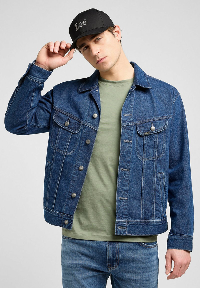 Denim jacket in dark blue with button front, two chest pockets, and a relaxed fit, worn over a light green shirt and paired with blue jeans.