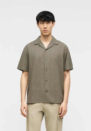 Jeune homme aux cheveux courts noirs portant une chemise boutonnée texturée à manches courtes de couleur olive et un pantalon cargo beige, debout devant un fond uni.