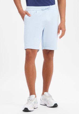 Mężczyzna stoi z rozluźnionymi rękami, ubrany w jasnoniebieskie szorty ze sznurkiem i białe buty sportowe na jednolitym tle.