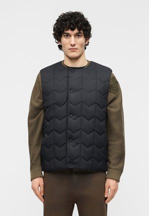 Ung mand med krøllet hår iført en sort quiltet vest over en tekstureret olivengrøn sweater, stående mod en ensfarvet baggrund.