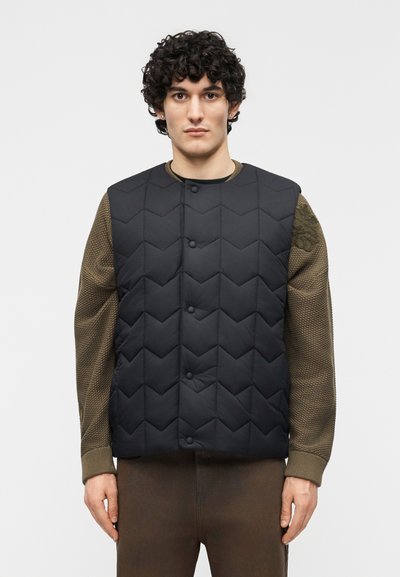 Jeune homme aux cheveux bouclés portant une veste matelassée noire sur un pull vert olive texturé, debout devant un fond uni.