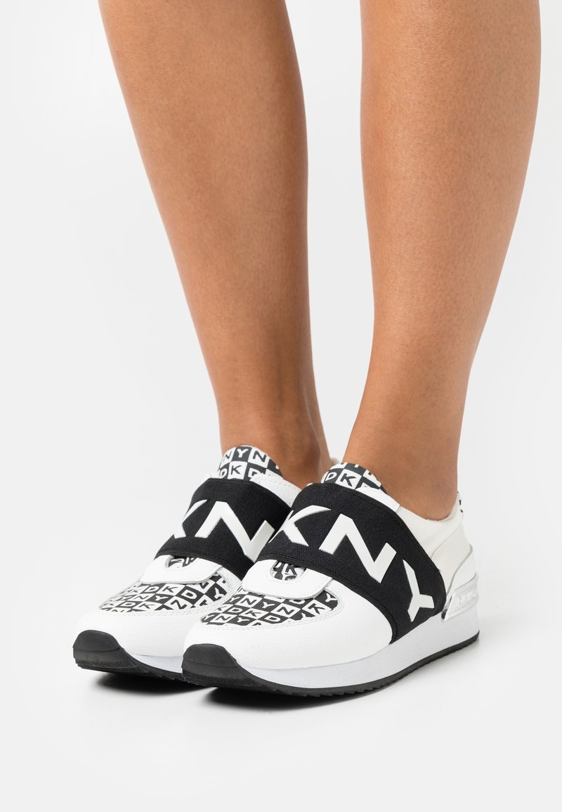 Weiße Sneaker mit schwarzen Akzenten, ausgestattet mit einem auffälligen Logo-Riemen und einem gemusterten Design im Obermaterial. Strukturierter Stoff und gepolsterte Sohle.