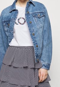 Džínová bunda s kovovými knoflíky, vrstvená sukňa s volány s květinovým vzorem a bílý tričko s květinovou aplikací.