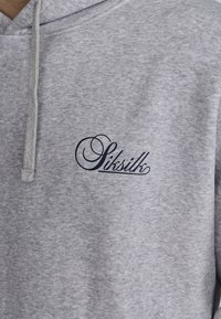 Pelēks kokvilnas kapucis ar priekšējo kangaroo kabatu, striķa kapuci un izšūtu "Siksilk" logo tumši zilā krāsā uz krūtīm.