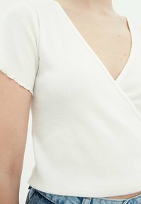 Top bianco a maniche corte con ruches e profondo scollo a V, con bordi delle maniche leggermente sfrangiati, abbinato a jeans di denim.