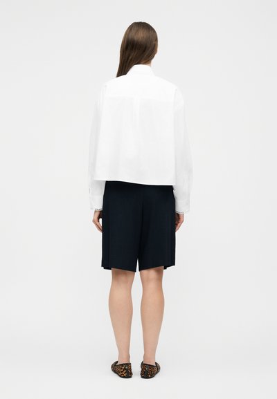 Femme aux longs cheveux bruns portant un haut blanc court à manches longues, un short noir arrivant aux genoux et des ballerines à imprimé léopard, debout de dos.