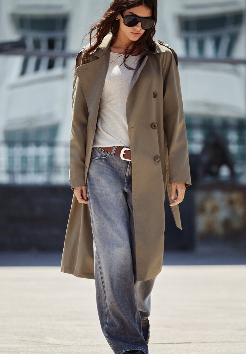 Femme portant des lunettes de soleil, un trench beige, un haut blanc et un jean bleu à jambes larges, marchant en extérieur dans un cadre urbain en journée.