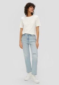 T-shirt bianco in cotone abbinato a jeans dritti chiari e strappati. Modella indossa sneakers bianche con plateau, in piedi su uno sfondo neutro.