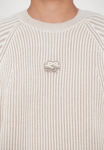 Crème en beige geribbelde gebreide trui met een klein geborduurd onderling knoopdesign op de borst, gedragen door een persoon.