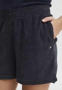 Svarte handklærshorts med elastisk midje, sidelommer og en liten metalllogo detalj på forsiden til høyre.