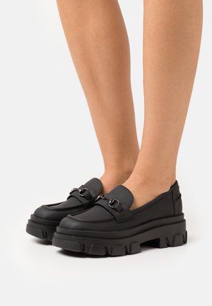 Mocassins noirs à semelle épaisse, fabriqués en matériau lisse. Présentent des détails en métal et un design épuré, sans motifs.