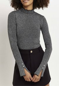 Grauer, gerippter Langarmoberteil mit einem Stehkragen und fransigen Bündchen, verziert mit dekorativen Knöpfen an den Ärmeln, kombiniert mit einem schwarzen Rock.