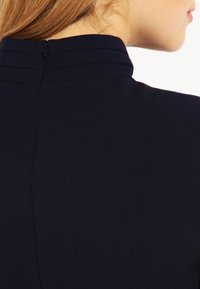 Granatowa sukienka z wysokim kołnierzem, subtelnymi pionowymi szwami, ukrytym zamkiem z tyłu i gładką fakturą.