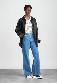 Melnas ādas pārējā izmēra krekls-jaka, balts svītrains polo krekls, gaiši zili platās kājas džinsi un melnbalti Sneakers. Gludas tekstūras, ikdienišķs izskats.