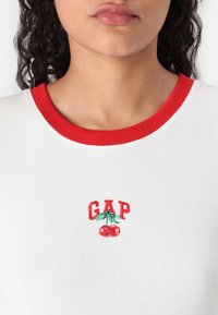 White ribbed T-shirt featuring a red ribbed neckline and an embroidered "GAP" logo with cherries beneath it.