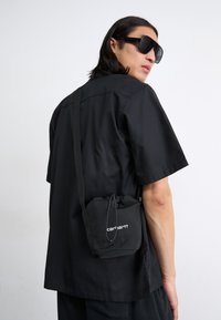 Black short-sleeve shirt worn by a person, paired with a small black drawstring bag featuring white logo. The setting is neutral and minimal.