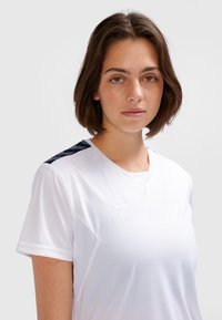 Biała sportowa koszulka z krótkimi rękawami, dekoltem w serek i teksturowanymi granatowymi akcentami na ramionach. Na piersi znajduje się wytłaczane logo.