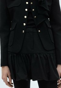 Costum stratificat negru, cu o jachetă ajustată și butoane aurii, și un tiv de fustă volanată, realizat dintr-un material neted cu un finisaj elegant.