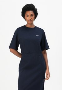 Ναυτικό μπλε βαμβακερό φόρεμα με κοντά μανίκια, στρογγυλή λαιμόκοψη και ελαστική μέση. Το λογότυπο της Barbour είναι κεντημένο στο στήθος.