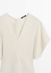 Beżowa sukienka z fakturowanej tkaniny, z krótkimi szerokimi rękawami, głębokim dekoltem w kształcie litery V oraz widocznym zamkiem błyskawicznym z tyłu na białym tle.