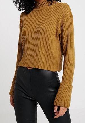 Femme portant un pull en maille côtelée moutarde avec des poignets évasés et un pantalon en cuir noir taille haute sur fond blanc.