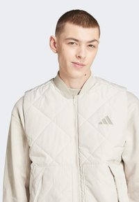 Chaleco acolchado de color beige claro con cierre de cremallera en el frente, cuello acanalado y dos bolsillos delanteros. Presenta un sutil logo gris de Adidas en el lado izquierdo del pecho.