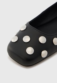 Chaussure plate en cuir noir avec un bout carré et ornée de petits clous ronds en argent sur la surface supérieure pour plus de détails.