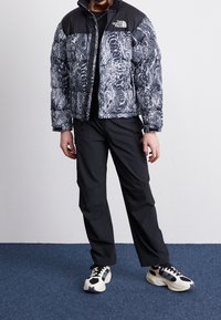 Pufferjakke med et mønstret gråt og sort krop, sort skulderområde og en frontlynlås. Kombineret med sorte bukser og sort-hvide sneakers.