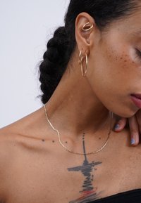 Gold-colored necklace with a wavy design resting on dark skin. Accessories include large hoop earrings and a cartilage hoop. Textured details are visible.