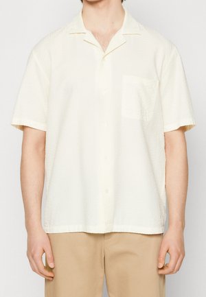 Man wearing short-sleeve, off-white textured button-up shirt with a chest pocket and beige pants, shown from shoulders to hips.