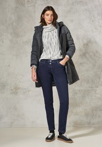 Doudoune grise avec des accents en polaire noire, pull à motifs blancs, jeans foncés et baskets noires avec des semelles marron, sur fond neutre.