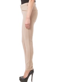Nainen, jolla on yllään beige slim-fit-housut, joissa on sivuvetoketju, ja mustat korot, seisoo sivuttain valkoista taustaa vasten.